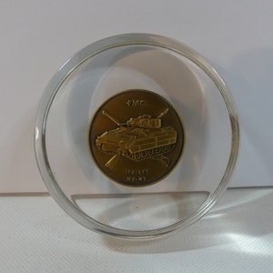 May include: A bronze-colored coin featuring a detailed illustration of a military tank, encased in a clear, circular acrylic holder. The coin has the text "FMC" at the top and "IFV-CFV M2-M3" at the bottom. The tank is the central focus.