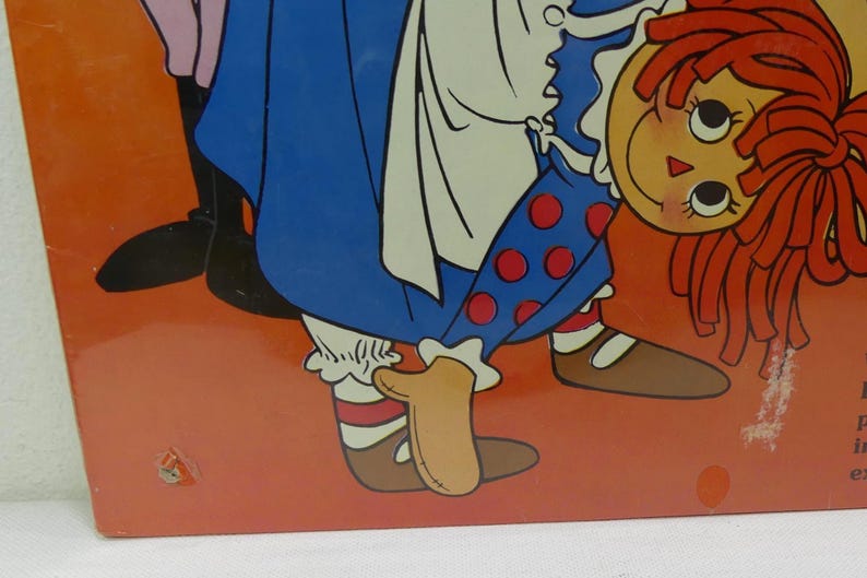 May include: A vintage illustration of a Raggedy Ann doll wearing a blue and white dress with red polka dots. She is standing on her hands with her legs in the air. The doll has red hair and a smiling face.