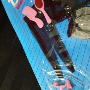 May include: A pink comb, a red and white hand mirror, and a pair of pink plastic shoes are packaged on a blue and white striped background. The packaging has the word "Dia" printed in pink letters.