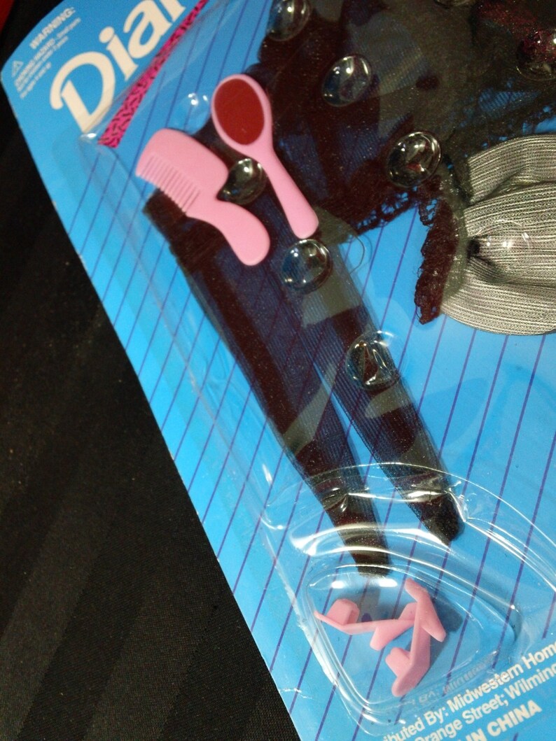 May include: A pink comb, a red and white hand mirror, and a pair of pink plastic shoes are packaged on a blue and white striped background. The packaging has the word "Dia" printed in pink letters.