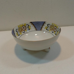 May include: A white footed bowl with a blue and yellow floral design. The bowl has a gold rim and three gold feet.