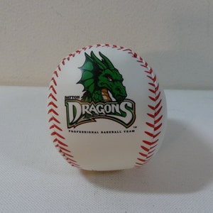 Puede incluir: Béisbol blanco con costuras rojas y un logotipo de dragón verde. El logotipo dice "Dayton Dragons" y "Professional Baseball Team".