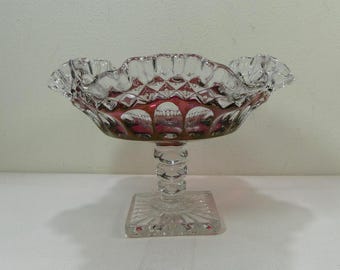 Puede incluir: Una compotera de cristal transparente con borde ondulado e interior rojo rubí. El cuenco tiene un diseño de diamantes y se apoya sobre un pie transparente y adornado y una base cuadrada. La compotera es probablemente vintage y decorativa.