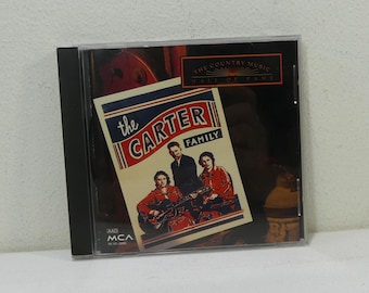 Puede incluir: Una caja de CD con la portada del álbum "The Carter Family". La portada muestra una fotografía vintage del grupo musical, con el texto "The Country Music Hall of Fame" en la parte superior. La caja del CD está en una funda de plástico transparente.