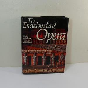 Puede incluir: El libro "La Enciclopedia de la Ópera" con una cubierta negra y una imagen colorida de un escenario con actores y estatuas. El título del libro está en texto blanco y el nombre del autor está en texto blanco más pequeño.