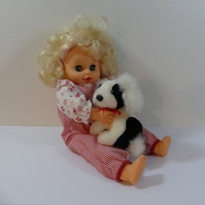 May include: A blonde doll wearing a red and white striped overall dress and a white shirt with red and white flowers. The doll is holding a black and white plush dog with a red bow.