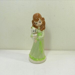 May include: A ceramic figurine of a girl in a light green dress holding a small white and green animal. The girl has brown hair and is standing on a white base. The figurine has a glossy finish and is likely vintage.