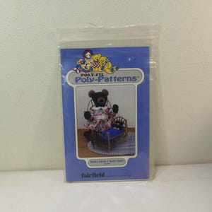 May include: A sealed plastic bag containing a blue and white pattern. The package features a graphic of a teddy bear wearing a floral dress and a small bed. The text "Poly-Fil Poly-Patterns" is visible.