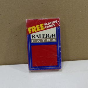 May include: A sealed deck of Raleigh Extra playing cards. The packaging is red with a blue border and the words "FREE PLAYING CARDS" in yellow and white. The brand name "RALEIGH EXTRA" is printed in white.