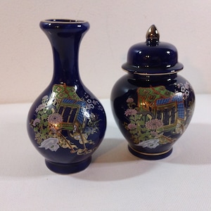 May include: Two navy blue ceramic vases. One is a slender vase, the other a lidded jar. Both are decorated with a colorful Asian-inspired design featuring buildings, flowers, and gold accents. The rims are trimmed with gold.