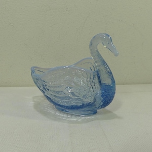 Può includere: Un piatto a forma di cigno in vetro blu chiaro. Il cigno ha un collo lungo e curvo e motivi di piume dettagliati sul corpo. Il piatto è un oggetto decorativo, probabilmente per l'esposizione o per contenere piccoli oggetti.