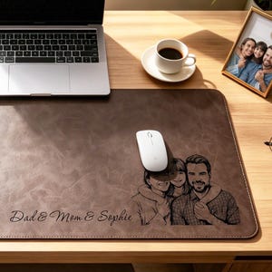 Op de afbeelding: Een bruine leren bureauonderlegger met een familiefoto en de tekst "Dad & Mom & Sophie" op een houten bureau. Een laptop, muis, koffiekopje en fotolijst staan ook op het bureau. Een gouden pennenhouder en een plant zijn zichtbaar.