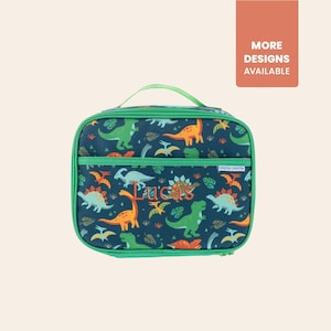 May include: A rectangular lunch bag with a green handle and trim. The bag features a navy blue background with colorful dinosaur illustrations. The name "Lucas" is embroidered on the front. The upper right corner has a peach-colored box with the text "MORE DESIGNS AVAILABLE."