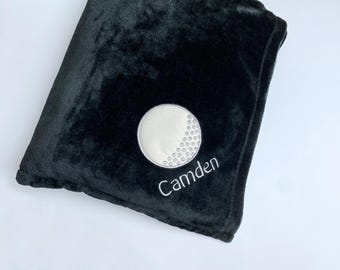 May include: Black plush blanket featuring an embroidered golf ball design. The golf ball is white with a silver outline and dimples. The name "Camden" is embroidered in white script below the golf ball.