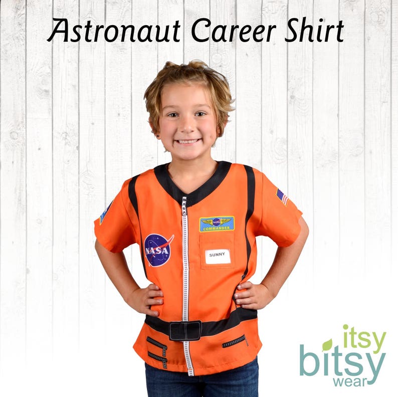 Puede incluir: Una persona joven lleva un traje de astronauta naranja con parches de la NASA y del Comandante. El traje tiene una cremallera en la parte delantera y un cintur&oacute;n negro. La persona sonr&iacute;e y tiene las manos en las caderas.
