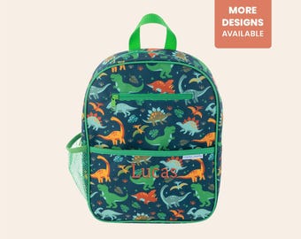 May include: A navy blue backpack with a green handle, trim, and zipper. The backpack features a colorful dinosaur pattern and the name "Lucas" embroidered in orange. A side pocket is made of green mesh. The text "MORE DESIGNS AVAILABLE" is in the upper right corner.