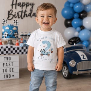 May include: A white short-sleeved t-shirt featuring a graphic of a blue race car, the number two, and the text "ITS MY BIRTHDAY" and "Harry". The design includes small balloons, stars, and checkered flags in blue, brown, and black colours.