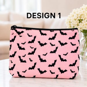May include: A rectangular zippered pouch in light pink featuring a repeating pattern of small black bats. The text DESIGN 1 is printed in black above the pouch. The pouch has a black zipper closure along the top edge and a flat base.