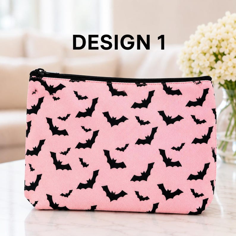 May include: A rectangular zippered pouch in light pink featuring a repeating pattern of small black bats. The text DESIGN 1 is printed in black above the pouch. The pouch has a black zipper closure along the top edge and a flat base.