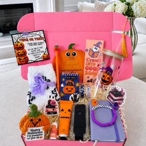Op de afbeelding: Een roze geschenkdoos gevuld met Halloween-artikelen, waaronder een pompoen-gezichtsmasker, een glazen beker met pompoenprint, een paars notitieboek, een gehaakte pompoen, pluizige witte sokken, een tube handcrème, een pen en een kaart met pompoenkunst.