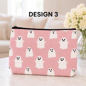 May include: A rectangular zippered pouch in a soft pink color featuring a repeating pattern of small white ghosts with black eyes and mouths. The text DESIGN 3 is printed in black above the pouch. The pouch has a black zipper closure.