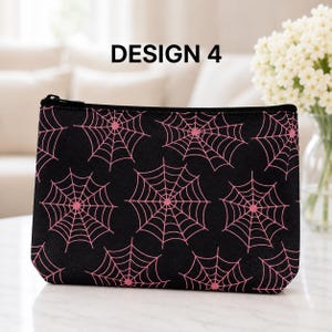 May include: A black rectangular zippered pouch featuring a repeating pattern of pink spider webs. The text DESIGN 4 is printed in black above the pouch. The pouch has a black zipper closure along the top edge and a structured, flat-bottomed shape.