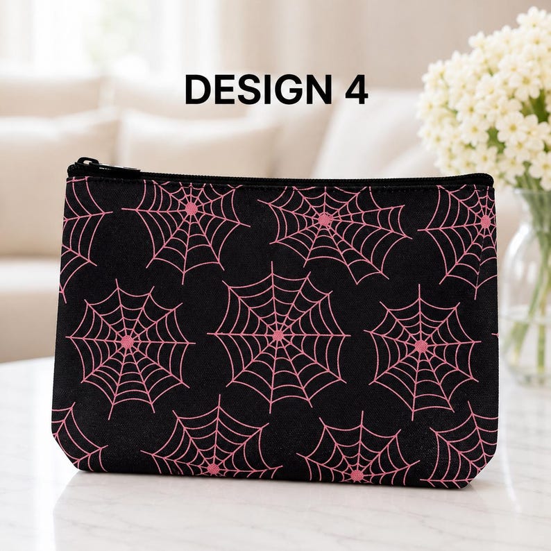 May include: A black rectangular zippered pouch featuring a repeating pattern of pink spider webs. The text DESIGN 4 is printed in black above the pouch. The pouch has a black zipper closure along the top edge and a structured, flat-bottomed shape.