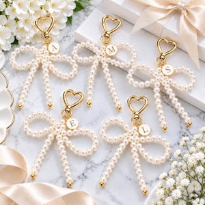 May include: Five gold-toned heart-shaped lobster clasp keychains, each featuring a bow made of white faux pearls. Each keychain includes a small circular charm with a single letter: A, M, S, E, or L. The keychains are arranged on a marble surface.