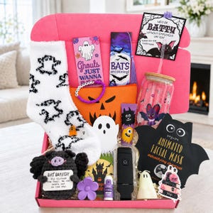 Può includere: Una scatola regalo rosa contenente articoli a tema Halloween: calzini bianchi soffici con motivi di fantasmi neri, un bicchiere di vetro con design a pipistrello, una maschera facciale nera a forma di pipistrello, un pipistrello all'uncinetto e carte.