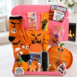 May include: A pink gift box filled with Halloween-themed items including orange fuzzy socks, a pumpkin-shaped face mask, a glass tumbler with a pumpkin print, a crochet pumpkin, a small notebook, a pen, a lip balm, and a card reading Trick or Treat.