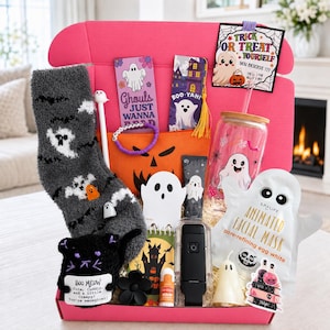 May include: A pink gift box filled with Halloween-themed items including a gray ghost-patterned sock, a glass tumbler with a ghost print, a SpaLife ghost-shaped facial mask, a black cat plush, bookmarks, a lip balm, and a ghost-themed card.