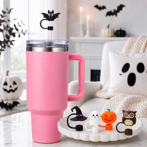 May include: A bright pink insulated tumbler with a handle and a clear lid, featuring a black bat-shaped straw topper. Beside it, a white scalloped plate holds four additional silicone straw toppers shaped like a witch hat, ghost, pumpkin, and owl.