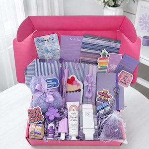 May include: A bright pink gift box filled with purple and blue themed items including a reading list card, a to-do list, a striped pencil case, a purple fuzzy blanket with a bow, a pen, a cupcake notepad, lotion, a book light, and stickers.