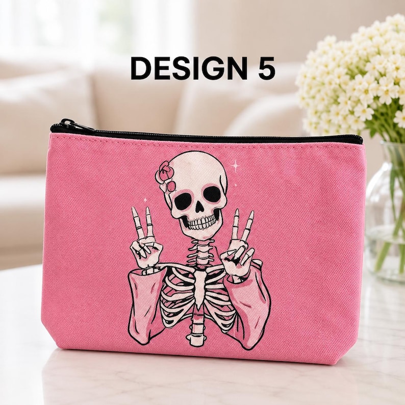 May include: A pink zippered pouch featuring a graphic illustration of a skeleton wearing a flower on its head and making peace signs with both hands. The text DESIGN 5 is printed in black above the pouch against a blurred background.