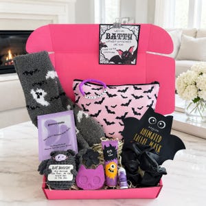 May include: A bright pink gift box containing gray fuzzy socks with white ghost patterns, a pink pouch with black bats, a purple eye mask, a crocheted bat toy, a purple skull bath bomb, a black flower clip, a monster-themed lotion, and a black scrunchie.