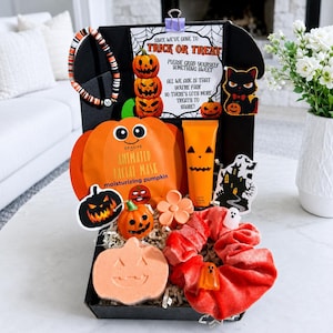 May include: A black gift box filled with Halloween-themed items including a pumpkin-shaped facial mask, an orange hand cream tube, a pumpkin bath bomb, a red velvet scrunchie with ghost charms, a beaded bracelet, and a sign reading Trick or Treat.