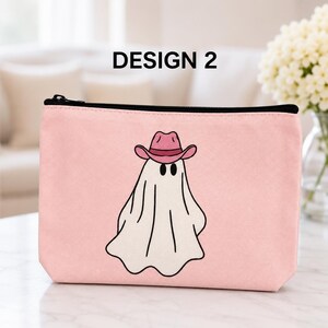 May include: A light pink rectangular zippered pouch featuring a central graphic of a white ghost wearing a pink cowboy hat. The ghost has simple black oval eyes and a black outline. The pouch has a black zipper closure along the top edge.