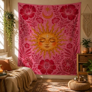 May include: Pink tapestry featuring a golden sun with closed eyes, surrounded by red flowers and foliage. A crescent moon and stars are above the sun. The tapestry is a decorative wall hanging.