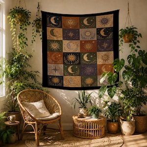 May include: A tapestry with a celestial design featuring suns, moons, and stars in a grid pattern. The tapestry is framed in black and hangs on a white wall. The room is decorated with plants, a wicker chair, and a small round table.
