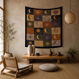 May include: A tapestry with a black border features a patchwork design of suns, moons, and stars in various colors. The squares are in shades of orange, green, purple, and beige. The tapestry is hanging on a neutral-colored wall.