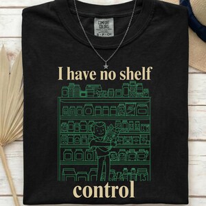 Könnte beinhalten: Ein schwarzes T-Shirt mit einer Grafik einer Person vor Regalen voller Gläser. Der Text "I have no shelf control" ist in cremefarbenen Buchstaben über und unter der grünen Strichzeichnung der Regale aufgedruckt.