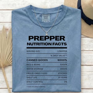 Könnte beinhalten: Ein blaues Kurzarm-T-Shirt mit schwarzem Text, der PREPPER NUTRITION FACTS lautet, und Elemente wie CANNED GOODS 9000%, RICE & BEANS 5000%, WATER STORAGE 3000%, FREEZE-DRIED FOOD STOCKED, SHELF LIFE 25 YEARS und SHELF CONTROL 0% auflistet.