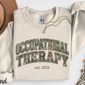 May include: A sand-colored crewneck sweatshirt featuring the text "OCCUPATIONAL THERAPY" in a camouflage print with a black outline, and the text "est. 2026" in brown below. A gold-toned star pendant necklace rests on the neckline of the garment.