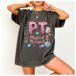 May include: Dark gray t-shirt with the letters "P.T." in pink hearts, and the words "Physical Therapist" below. The shirt features illustrations of exercise equipment, a knee joint, and crutches.