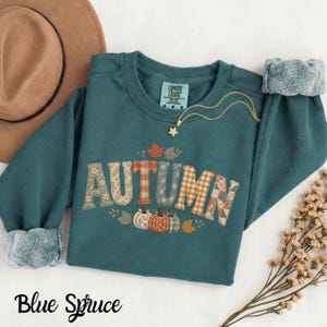 Könnte beinhalten: Ein blaugrünes Sweatshirt mit Rundhalsausschnitt und dem Wort AUTUMN in großen, gemusterten Blockbuchstaben mit Blumen- und Karodesigns. Unter dem Text befinden sich drei kleine Kürbisse in Orange, Weiß und Grün. Eine goldene Sternkette liegt am Kragen.