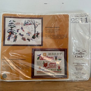 May include: A package containing two cross-stitch kits from The Creative Circle. The kits are titled "2310 Snow Angels" and "2311 Messages of Christmas". The Snow Angels kit features a winter scene with children playing in the snow. The Messages of Christmas kit features a colourful design with the words "Merry Christmas" and "Peace on Earth".
