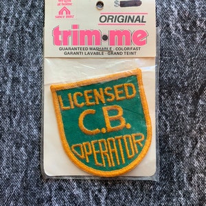 May include: A green and gold embroidered patch with the text "LICENSED C.B. OPERATOR" is displayed in a clear plastic package. The patch is shield-shaped. The packaging reads "trim-me" and "ORIGINAL".