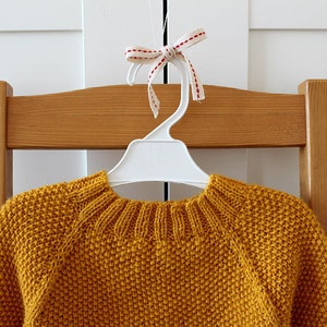 May include: A mustard yellow knitted sweater hangs on a white plastic hanger with a red and white ribbon bow. The sweater has a round neckline and long sleeves.
