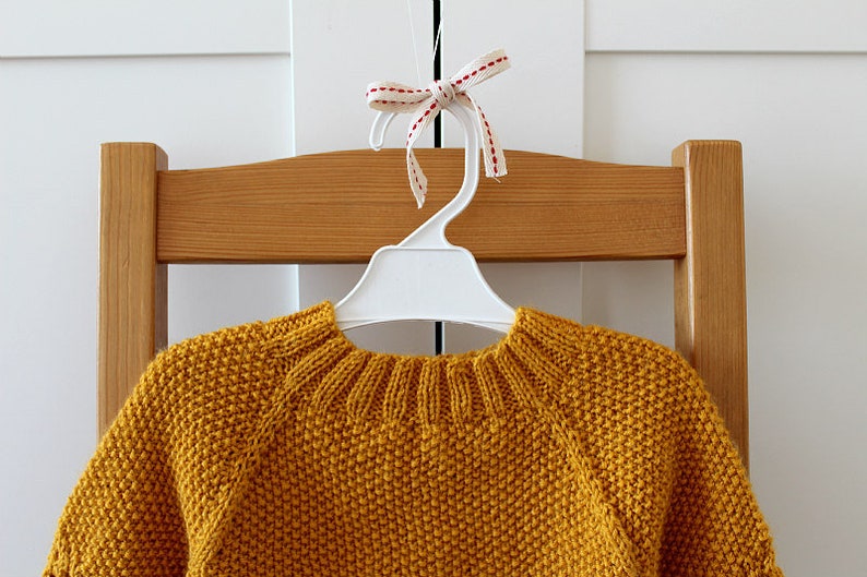 May include: A mustard yellow knitted sweater hangs on a white plastic hanger with a red and white ribbon bow. The sweater has a round neckline and long sleeves.