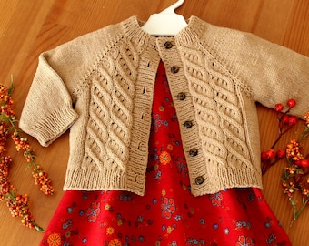 May include: A beige knitted cardigan with a cable knit pattern and button closure. The cardigan is worn over a red dress with a floral print.